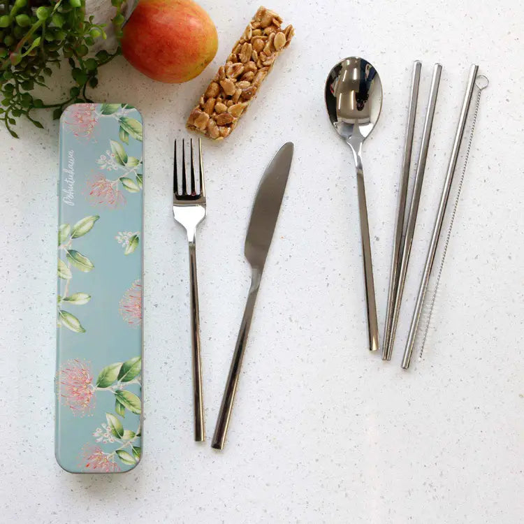 A flat-lay of a stainless steel cutlery set with a floral tin case on a white speckled countertop. The set includes a fork, knife, spoon, two metal chopsticks, and two straw-like utensils, alongside a peanut butter bar and greenery as props.