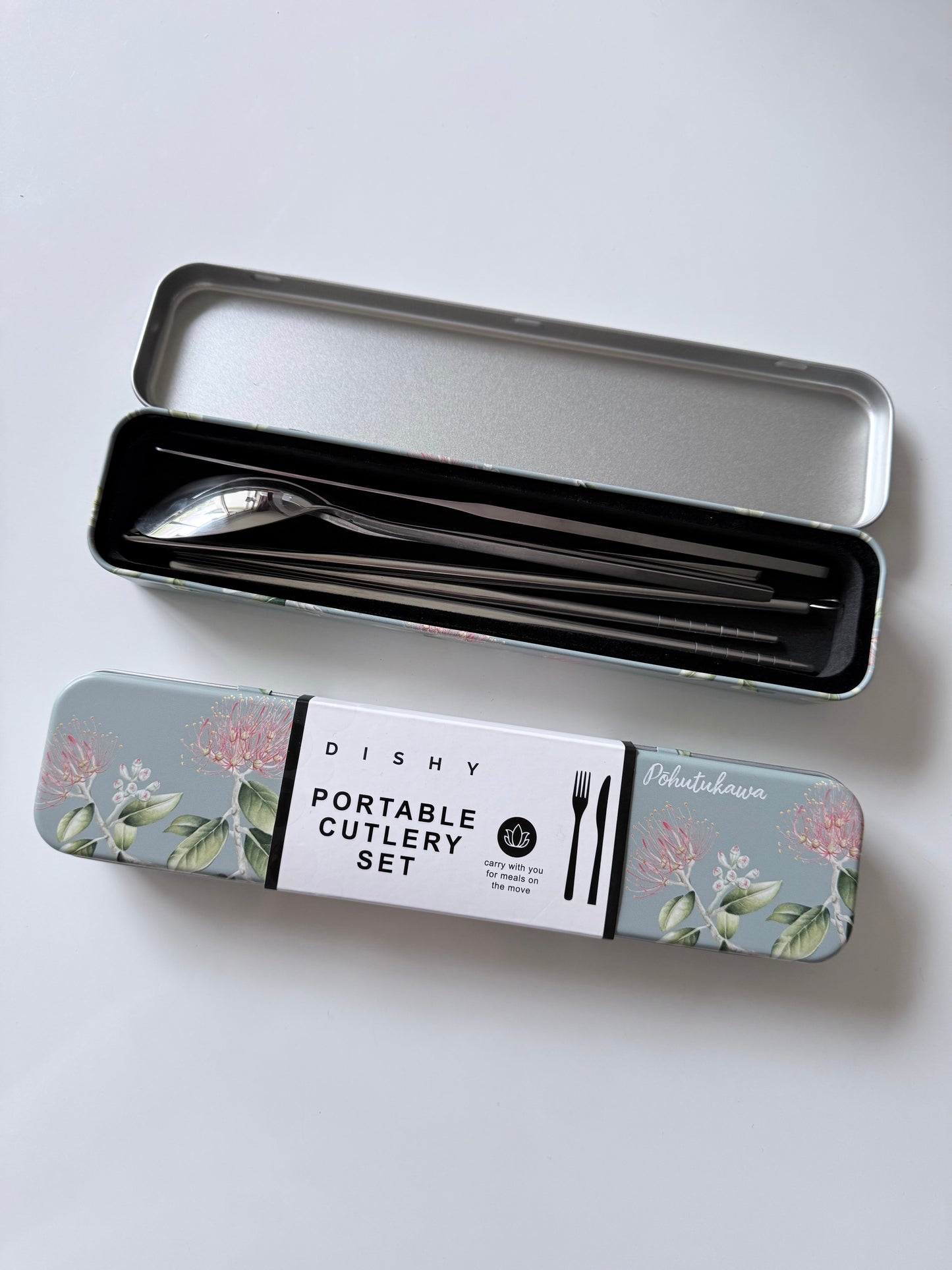 A portable cutlery set in a rectangular metal tin with floral artwork on the lid, shown open with utensils inside and the closed lid placed below it on a light gray background.