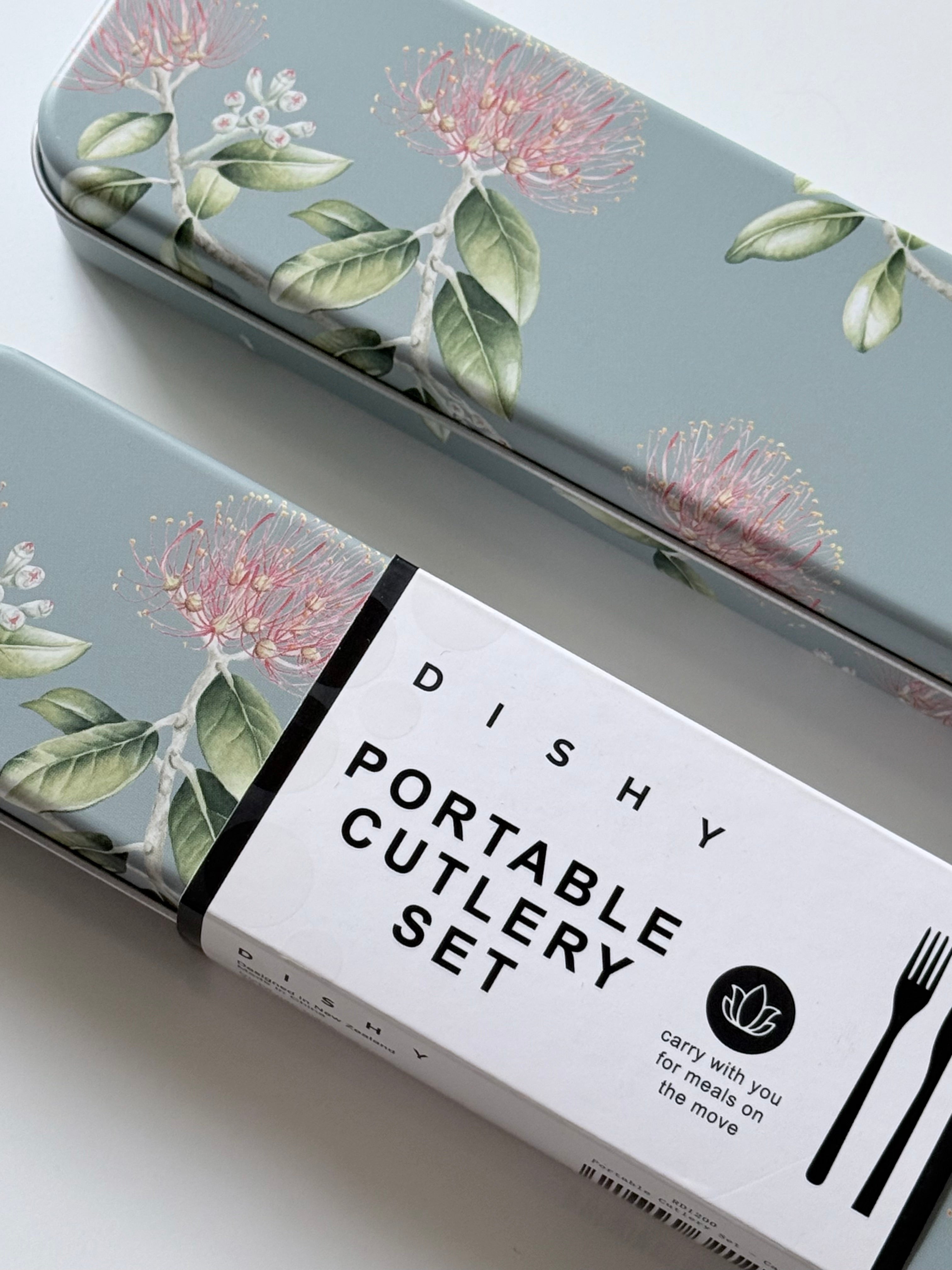 A portable cutlery set in a rectangular metal tin with a muted blue-gray background and a botanical print of pink flowers and green leaves. A white paper band around the tin reads 'DISHY PORTABLE CUTLERY SET' with a small leaf logo and black utensil silhouettes.